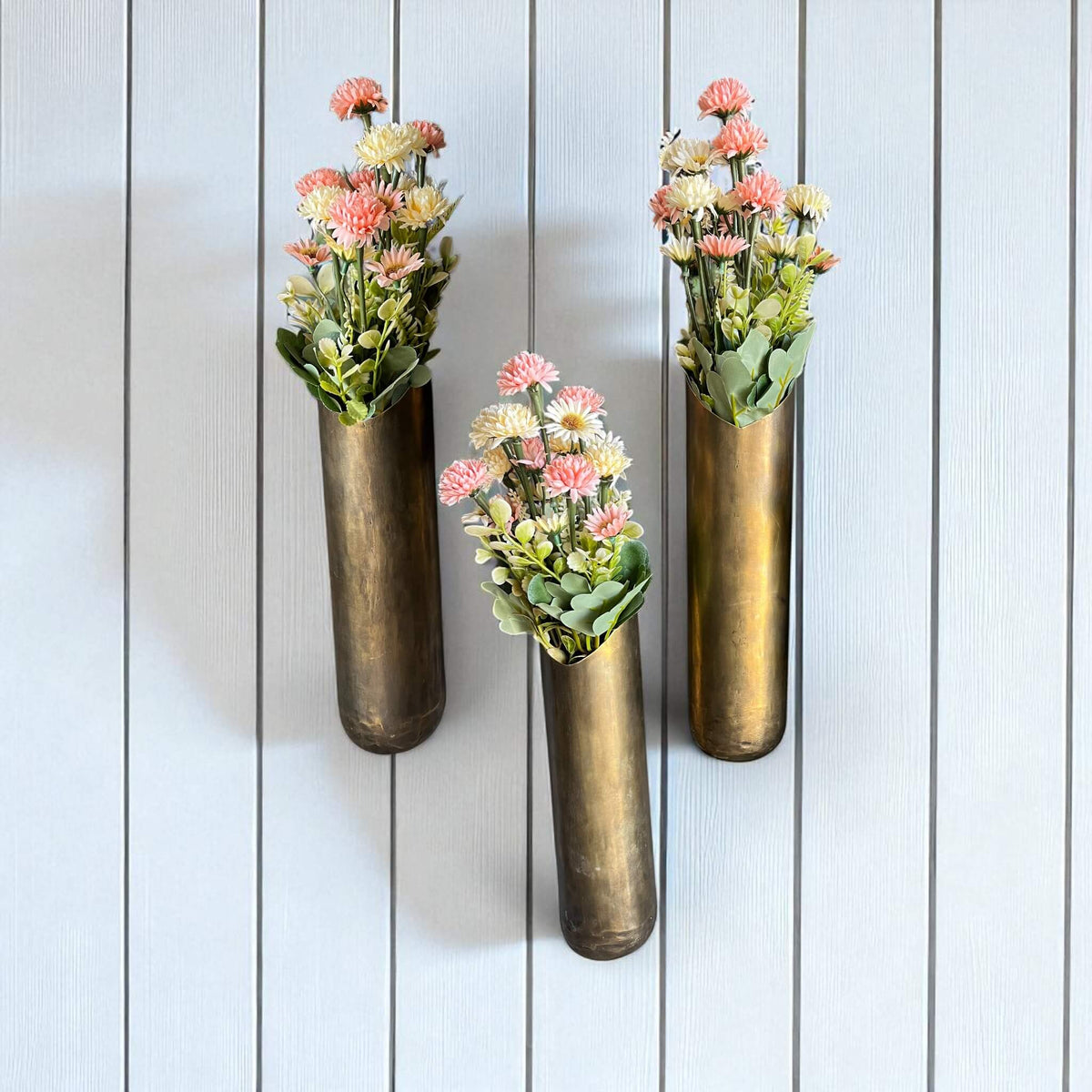 Three brass wall vases with floral arrangements on a light wooden panel background