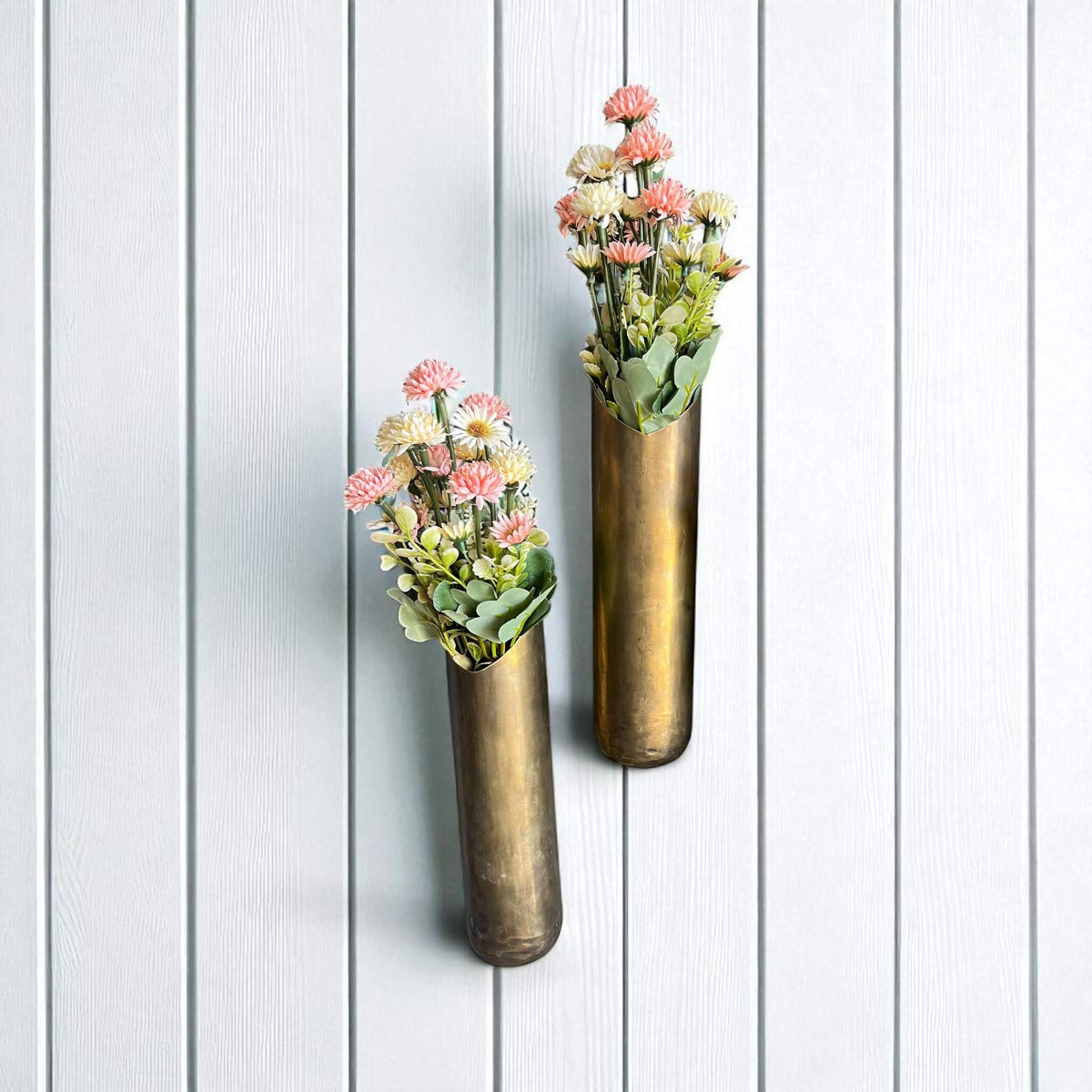 Two brass wall vases with floral arrangements on a white wooden background